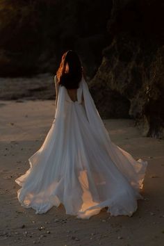 a woman in a white wedding dress standing on the beach with her back to the camera