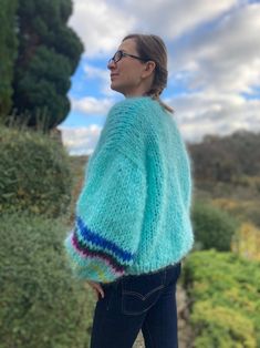 a woman standing in front of some bushes wearing a blue sweater and black jeans with her hands on her hips