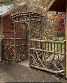 a wooden gate made out of branches in front of a brown building with trees around it