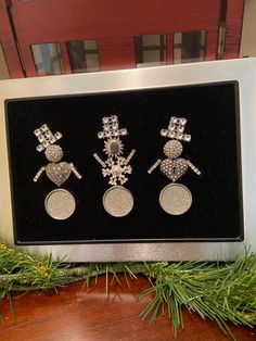 three snowman earrings are on display in a box with pine branches and evergreens