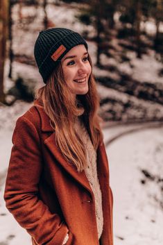 a woman standing in the snow wearing a brown coat and a black beanie hat