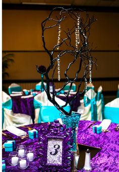 the table is set up with purple and blue linens, white candles, and a tree branch centerpiece