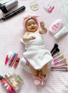 a baby laying on top of a pink blanket next to makeup and other cosmetics items