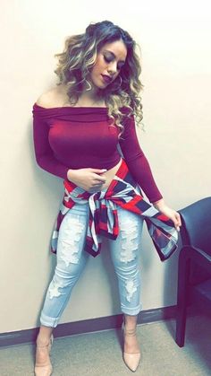 a woman leaning against a wall wearing ripped jeans and a red off the shoulder top