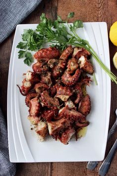 an octopus dish on a white plate with lemons and parsley next to it