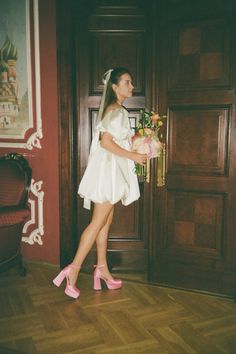 a woman in a white dress and pink high heels is standing by a door holding a bouquet of flowers