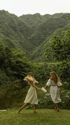 two women in white dresses are holding hands by the water's edge with mountains in the background