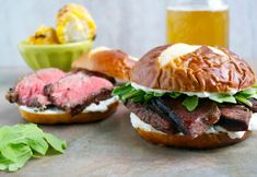 two steak sandwiches with lettuce, cheese and other toppings next to a glass of beer