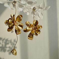 a pair of gold and white flower earrings hanging from a branch with leaves on it