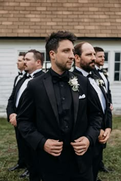 a group of men standing next to each other in front of a white building and wearing black suits