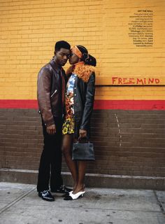 a man and woman standing next to each other in front of a yellow brick wall