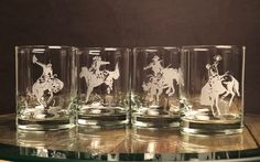 four glasses with etched cowboy images on them sitting on a table in front of a brown wall