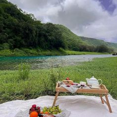 a picnic table with fruit and tea on it next to a river in the mountains