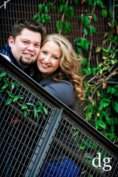 a man and woman standing next to each other in front of a fence with green leaves