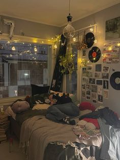 an unmade bed with lots of clothes on it and lights hanging from the ceiling