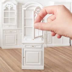 a hand is holding the door to a dollhouse kitchen with white cabinets and cupboards