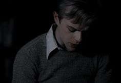 a man in a dark room looking down at his cell phone while wearing a sweater