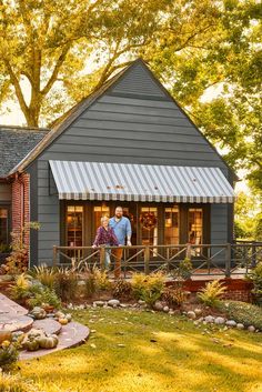 two people standing on the front porch of a small gray house with an awning