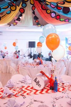 balloons and streamers fill the ceiling at a party with white tablecloths, red and orange striped tables