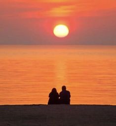 two people are sitting on the beach watching the sun go down over the water at sunset