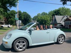 four people in a light blue convertible driving down the street with trees and houses behind them