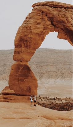 two people standing in front of an arch shaped rock formation on the side of a desert