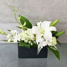 a black vase with white flowers and green leaves sitting on a glass table in front of a gray wall