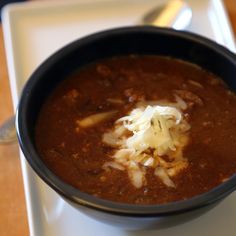 a black bowl filled with chili and cheese on top of a white countertop next to a spoon