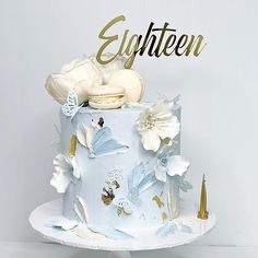 a blue cake with white flowers and gold lettering that says eyghteen