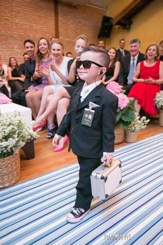 a young boy dressed in a suit and sunglasses carrying a suitcase while people look on