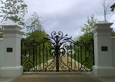 an iron gate with decorative designs on the top and bottom bars, in front of a driveway