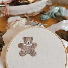 a teddy bear is embroidered onto a white piece of fabric in front of a basket full of yarn