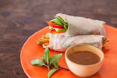 an orange plate topped with two rolls and dipping sauce on top of each other next to a wooden bowl