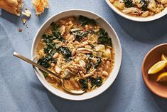 two bowls of soup with spinach, chicken and lemon slices next to bread on a blue surface