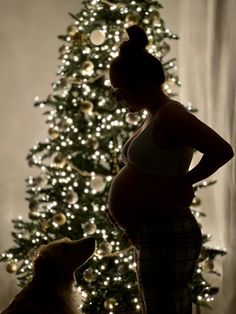 a pregnant woman standing in front of a christmas tree with her dog looking at it