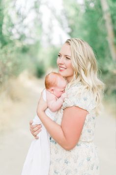 a woman holding a baby in her arms and smiling at the camera with trees in the background