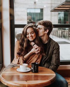 a man and woman sitting at a table in front of a window with their arms around each other