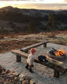 a little boy standing next to a fire pit on top of a wooden bench in the middle of a field