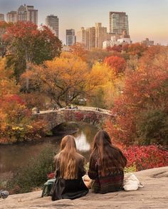 two women sitting on the edge of a cliff overlooking a river and bridge in autumn