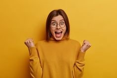 a woman with glasses and an excited look on her face, standing in front of a yellow background