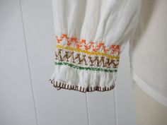 "Vintage 1970s peasant embroidered shirt, ethnic blouse. Made of white cotton. Has orange, yellow and brown squash blossom detail. Maybe Navajo. String tie at the neck. No label. Actual measurements are: 39\" at the bust 39\" at the waist 17\" shoulder to shoulder 19\" shoulder to end of sleeve 23\" overall length In very good condition with a small faint spot(photo)." 1970s Blouse, Peasant Shirt, Hippie Shirt, Squash Blossom, Vintage Blouse, Blouse Vintage, Embroidered Shirt, Yellow And Brown, Vintage 1970s