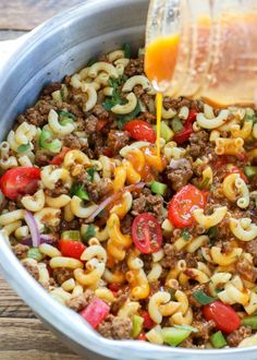 a person pouring dressing into a bowl filled with pasta and ground beef, tomatoes, bell peppers, green onions, red onion, carrots, celery lettuce