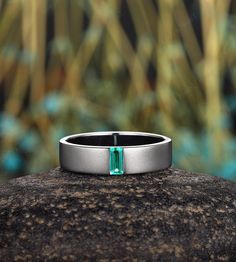 a wedding band with an emerald colored stone in the center on top of a rock