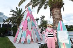 a woman standing next to a pink and blue teepeed tent on top of a palm tree