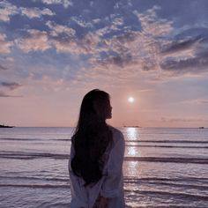 a woman standing on the beach looking at the ocean and setting sun in the distance