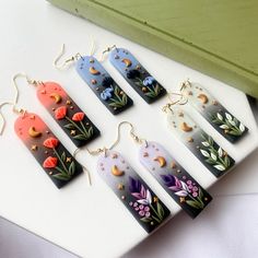 five pairs of earrings with flowers painted on them sitting on a white surface next to a green box