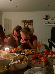 three women sitting at a table with food and drinks in front of them, while one woman holds her face to her mouth