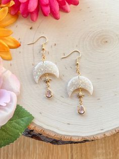 a pair of earrings sitting on top of a piece of wood next to pink flowers