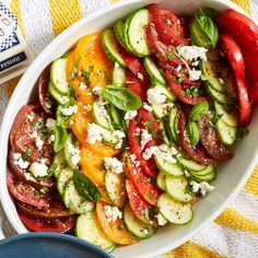 a white bowl filled with cucumbers, tomatoes and other veggies next to a bottle of feta cheese