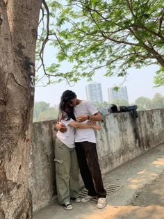 two people standing next to each other near a tree and cement wall with buildings in the background